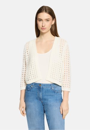 Ung kvinde med lige brunt hår iført en hvid åbenstrikket cardigan over en hvid top og blå højtaljede jeans, stående foran en neutral baggrund.