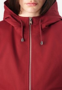 Kvinne iført en hettejakke i dyp rød med glidelås og snorer med sølvfargede metalltupper.