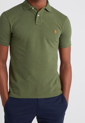 Mann trägt olivgrünes kurzärmeliges Poloshirt mit orangefarbenem Logo auf der Brust und dunkelblaue Hose, Hände in den Taschen, vor einfarbigem Hintergrund.