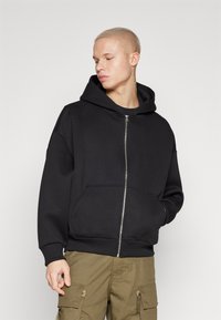 Black zip-up hoodie, made of fleece, features a large hood, front pockets, and ribbed cuffs. Worn with green cargo pants.
