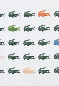 Λευκό ύφασμα με επαναλαμβανόμενο μοτίβο κροκόδειλων σε πράσινο, πορτοκαλί, ροζ, μπλε και πράσινο. Υφασμάτινη υφή στις λεπτομέρειες των κροκόδειλων.