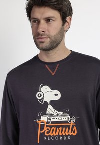 Camiseta de manga larga gris oscuro con un gráfico de Snoopy usando auriculares y una mesa de mezclas. Costuras naranjas destacan en el cuello.