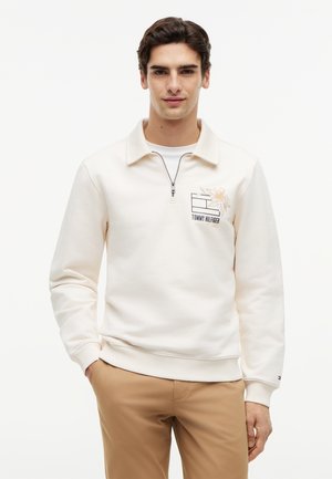 Młody mężczyzna ubrany w kremową bluzę z suwakiem do ćwiartki, ozdobioną kwiatowym wzorem i logo na piersi, w połączeniu z beżowymi spodniami, stojący na białym tle.