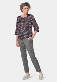 Blumenbluse in Schwarz mit lila und grünen Mustern; Dreiviertelärmel. Graue, taillierte Hose; weiße Sneakers mit hellgrauen Akzenten.