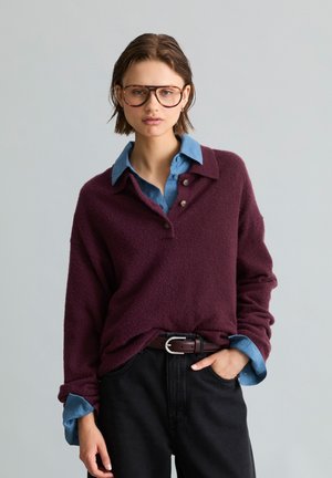 Junge Frau mit kurzen Haaren, die große Brille trägt, einen burgunderfarbenen Pullover über einem blauen Hemd mit Kragen, schwarze Hose und Gürtel, posiert mit einer Hand in der Tasche.