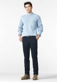 Lichtblauwe gebreide trui met een voorzak, gedragen met donkerblauwe broek en beige laarzen, staand tegen een eenvoudige witte achtergrond.