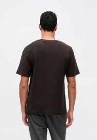 Muž s krátkymi kučeravými vlasmi, oblečený v jednoduchom tmavohnedom tričku s krátkym rukávom a tmavých nohaviciach, z pohľadu zozadu na bielom pozadí.