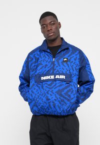 Blå pulloverjakke har et geometrisk mønster, justerbar krave, brystlomme med "NIKE AIR" logo og elastiske manchetter og nederkant.
