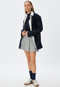 Mørkeblå blazer over en hvid skjorte, parret med en tekstureret grå nederdel, mørkeblå strømper og hvide sneakers med sorte accenter. En afslappet og lagdelt stil.