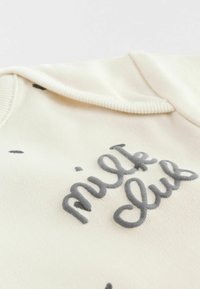 Valkoinen huppari, jossa on pehmeästi harmaalla brodeerattu "milk club" -teksti. Sileä kangas, jossa on ribbiyhdiste ja satunnaisia mustia pisaroita.