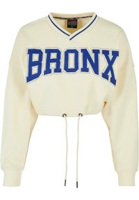 Cropped cream sweatshirt with blue "BRONX" text in large letters, embellished with rhinestones. Features a V-neck and adjustable drawstring hem.