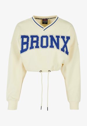 Sutrumpinta kreminė sportinė palaidinė su mėlynu dideliu tekstu "BRONX", puošta cirkoniais. Turi V formos iškirptę ir reguliuojamą gaubtą.