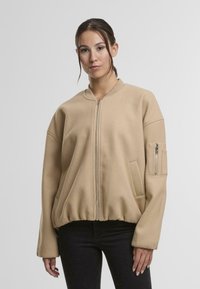 Beige zipovací bunda vyrobená z měkkého materiálu, s žebrovaným lemem, oversized střihem a zipovou kapsou na levém rukávu.