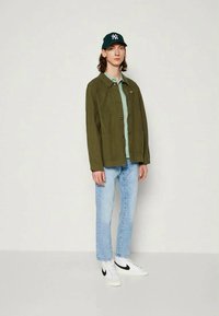 Olivgrön overshirt med knappar, ljusblå t-shirt, ljusblå denimjeans och vita sneakers med svarta accenter samt en grön keps.