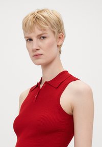 Rød ribbestrikket ermeløs polo-overdel med krage, med knappestolpe og tettsittende design som fremhever en jevn tekstur og rene linjer.