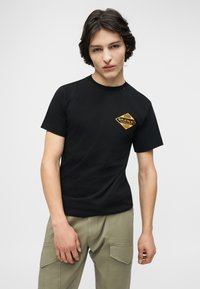 Νεαρός άνδρας με μεσαίου μήκους σκούρα μαλλιά, φορώντας μαύρο μπλουζάκι με κίτρινο λογότυπο "Miami Replay Palace Florida" και χακί παντελόνι cargo.