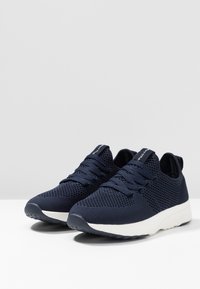 Granatowe siatkowe sneakersy o strukturalnej powierzchni, z sznurowanym zapięciem i białą podeszwą środkową; posiadają pętelkę z tyłu ułatwiającą zakładanie.