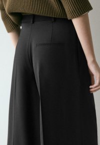 Schwarze, weit geschnittene Hose mit glatter Textur, ausgestattet mit einer Gesäßtasche und einem maßgeschneiderten Bund. Der Stoff wirkt leicht und fließend.