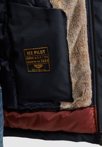 Veste marine avec un col en fourrure, poche latérale avec une étiquette jaune et doublure intérieure rouge. Les textures notables incluent un tissu lisse et une fourrure douce.