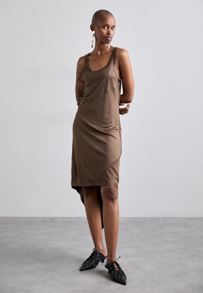 Robe sans manches marron avec un motif géométrique. Tissu doux et extensible, avec un ourlet asymétrique. Portée avec des chaussures noires à détails froncés.