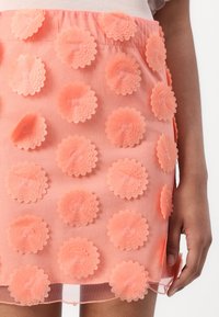 Pupurrete skjørt laget av peach-farget stoff, med lagdelte blomst-appliquéer med sirkulære kronblad og små midtperler, glatt tekstur og over-kneelengde.