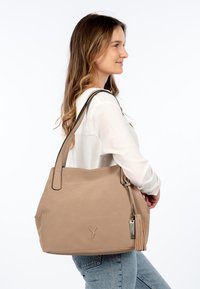Beige textured hobo bag with black accents, shoulder strap, and a tassel detail. Features a logo embossed on the front.