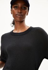 Schwarzer gerippter Strickpullover mit kurzen Ärmeln und weitem Ausschnitt, kombiniert mit einer marineblauen Kappe. Das Material wirkt weich und strukturiert.