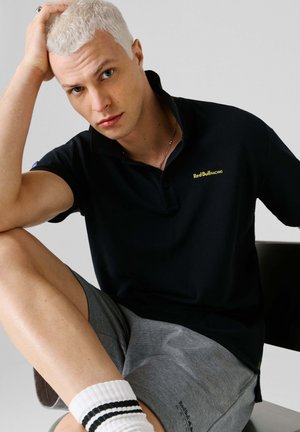 Noor blondide juustega noormees, kes kannab musta Red Bull Racing polo, hallid lühikesed püksid, triibulised valged sokid, istub ja toetab pead käega.