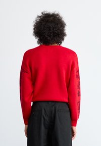 Suéter de punto rojo con mangas largas que presenta diseños gráficos en negro en las mangas; ajuste holgado y cuello redondo. Se aprecian pantalones negros por debajo.