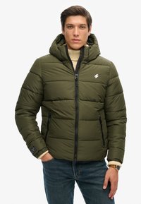 Polstret grøn jakke med hætte, lynlås lukning og to sidelommer. Har et quilted mønster og et lille logo på brystet.