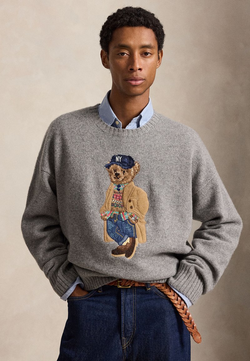 Jeune homme portant un pull gris avec un ours en peluche portant un manteau, un pull, un jean et une casquette NY, associé à une chemise bleue et une ceinture marron tressée.