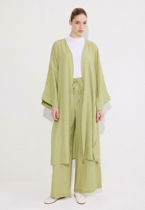 Naine, kes kannab lahtist helerohelist avatud kardigani ja sobivaid laia säärega pükse valge topi ja valgete kingadega, seisab ühevärvse tausta ees.