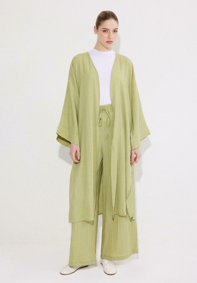 Γυναίκα που φορά χαλαρό ανοιχτό πράσινο ζακέτα και ταιριαστό παντελόνι με φαρδιά μπατζάκια πάνω από λευκό τοπ με λευκά παπούτσια, στέκεται μπροστά σε απλό φόντο.