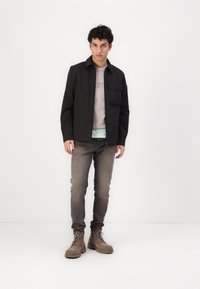 Sort jakke med krave og frontlomme, lagdelt over en grå skjorte og en lyseblå undertrøje, parret med grå jeans og brune støvler.