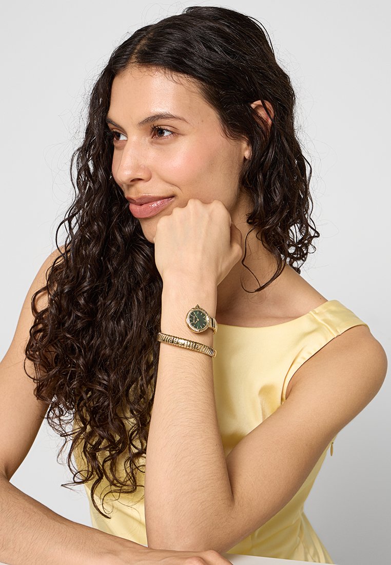 Orologio da polso dorato con quadrante verde e cassa rotonda, abbinato a un bracciale in oro testurizzato, indossato su un braccio di pelle chiara su uno sfondo neutro.