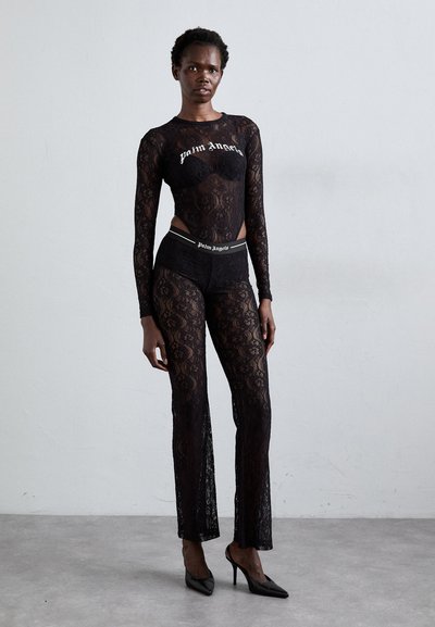 Black lace long-sleeve top and matching high-waisted pants with a logo waistband. Features sheer texture and fitted design. Heels complete the look.