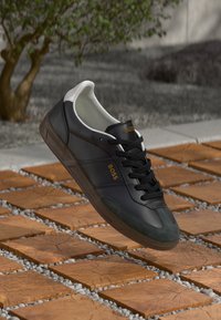 Baskets en cuir noir avec lacets noirs et contrefort de talon blanc, marquées "BOSS" en doré, flottant au-dessus d'un sol en carreaux de bois et galets.