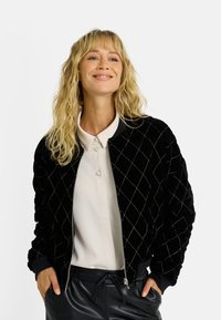 Blouson bomber en velours noir avec motif matelassé diamant, fermeture éclair à l'avant, poignets côtelés et col lisse. Porté sur un chemisier léger.