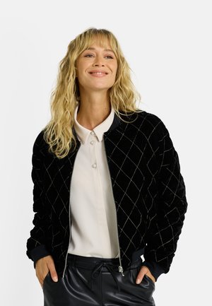 Chaqueta bomber de terciopelo negro con patrón acolchado en forma de diamante, cierre frontal con cremallera, puños acanalados y cuello liso. Llevada sobre una blusa clara.