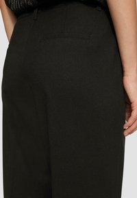 Pantalons noirs ajustés avec une texture de tissu lisse, des détails subtils de poche et une coupe droite. Main positionnée près de la taille.