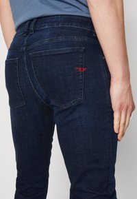 Jeans de mezclilla azul oscuro con corte ajustado, que cuentan con dos bolsillos traseros y costuras rojas en el bolsillo derecho. Textura suave y diseño clásico.