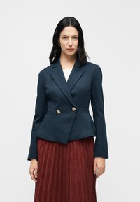 Blazer bleu marine à design péplum avec des boutons floraux dorés, assorti à une jupe plissée bordeaux. Texture lisse et coupe ajustée.