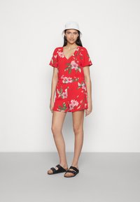 Macacão floral vermelho com decote em V, mangas curtas e cintura elástica. Realçado com flores cor-de-rosa e folhas verdes, combinado com sandálias pretas.