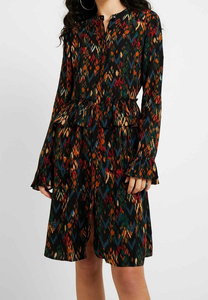 Femme portant une robe à manches longues avec un motif multicolore, taille et poignets à volants, debout devant un fond uni.