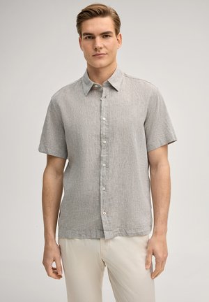 Junger Mann mit kurzen Haaren, der ein hellgraues kurzärmliges Hemd mit Knöpfen und beige Hosen trägt, steht vor einem einfarbigen Hintergrund.