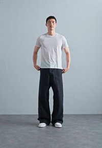 White cotton t-shirt paired with loose-fitting, solid black pants and white sneakers. Model stands against a plain gray background.