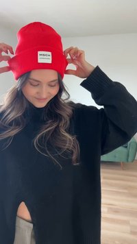 Berretto rosso lavorato a maglia con etichetta, indossato da una persona in un maglione extra-large nero, con lunghi capelli ondulati e un ambiente domestico rilassato.