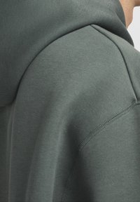 Kuklová mikina z tmavě zelené bavlněné tkaniny. Obsahuje zaoblený ramenní šev a hladkou texturu. Žádné viditelné vzory ani kovové prvky.