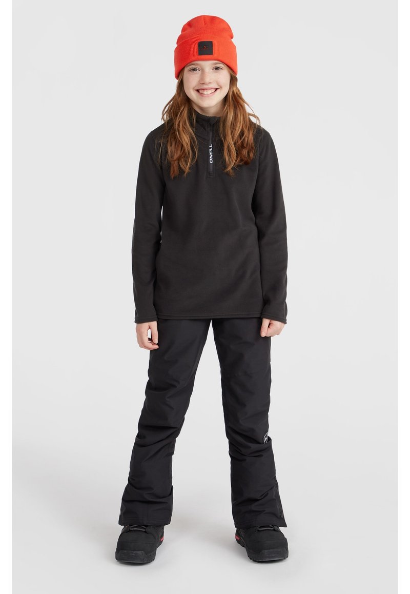 Pullover in pile nero con colletto con zip, abbinato a pantaloni da neve neri. Il modello indossa un berretto arancione acceso e stivali neri.
