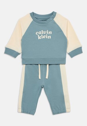 Dwuczęściowy, niebiesko-kremowy strój dla niemowlaka z długimi rękawami, elastycznymi mankietami, spodniami ze sznurkiem i napisem "calvin klein" na piersi.